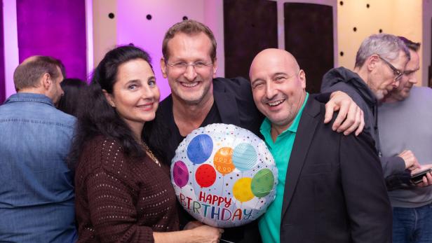 Drei lachende Menschen halten einen Geburtstagsballon mit bunten Luftballons und der Aufschrift „Happy Birthday“.