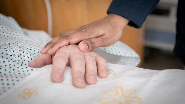 Eine Hand liegt tröstend auf einer anderen Hand, die auf einer Bettdecke mit Blumenmuster ruht.