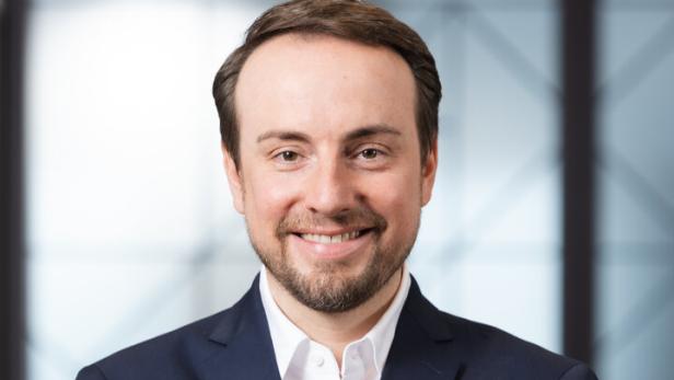 Ein Mann mit dunklem Sakko und weißem Hemd steht mit verschränkten Armen und lächelt freundlich.