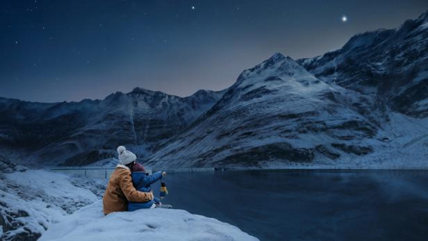 Zwei Personen sitzen am verschneiten Ufer eines Stausees vor schneebedeckten Bergen bei Nacht.