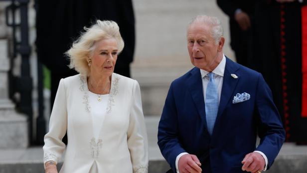 König Charles und Königin Camilla schreiten nebeneinander, er im blauen Anzug, sie in einem weißen Kleid.