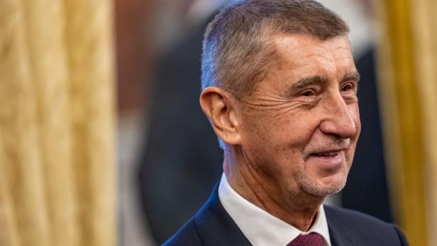 Ein älterer Mann im Anzug mit roter Krawatte lächelt und steht vor einem unscharfen Hintergrund mit goldenen Vorhängen.