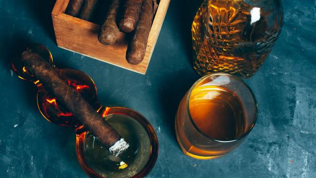 Eine brennende Zigarre liegt in einem Aschenbecher neben einem Glas und einer Karaffe mit braunem Getränk sowie einer Zigarrenkiste.