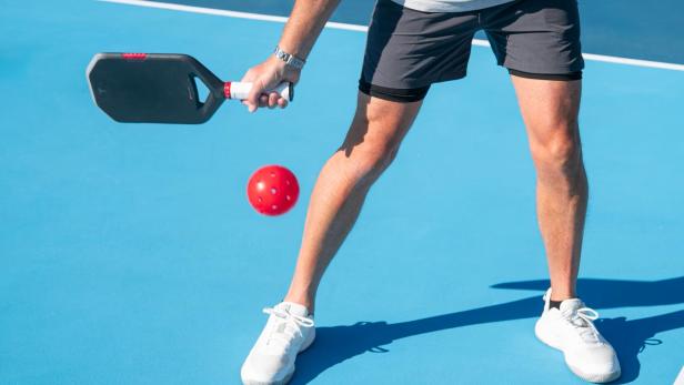 Person spielt Pickleball mit schwarzem Schläger und rotem Ball auf blauem Spielfeld.
