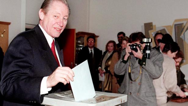 Ein Mann im Anzug wirft einen Wahlzettel in eine Wahlurne, während Fotografen die Szene festhalten.