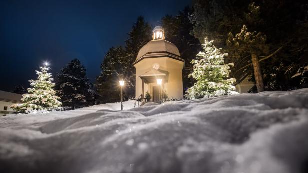 Verschneite kleine Kapelle bei Nacht, umgeben von beleuchteten Tannenbäumen und Laternen im Schnee.