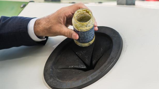 Eine männliche Hand wirft ein leeres Pestoglas in einen Glascontainer.