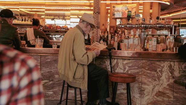 Ein älterer Mann mit Hut sitzt an einer Bar, liest eine Zeitung und hat einen Gehstock neben sich abgestellt.