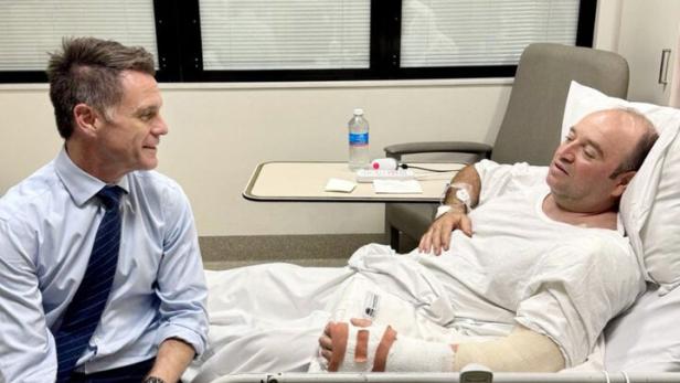 Ein Mann im Anzug sitzt am Krankenbett eines verletzten Mannes