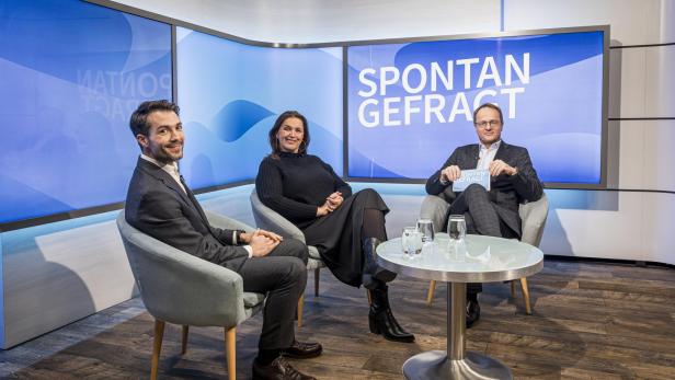 Drei Personen sitzen in Sesseln um einen runden Tisch mit Gläsern und Wasserkanne, im Hintergrund Bildschirm mit Schriftzug 'SPONTAN GEFRACT'.