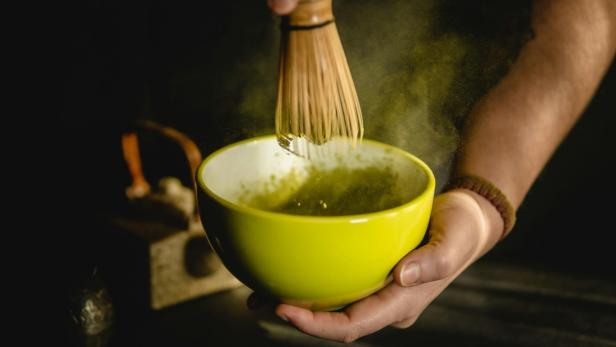 Hand hält eine gelbe Schale, in der mit einem Bambusbesen grünes Pulver aufgeschlagen wird.