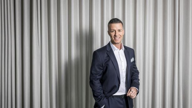 Ein Mann im dunklen Anzug mit weißem Hemd steht lächelnd vor einem hellgrauen Vorhang.