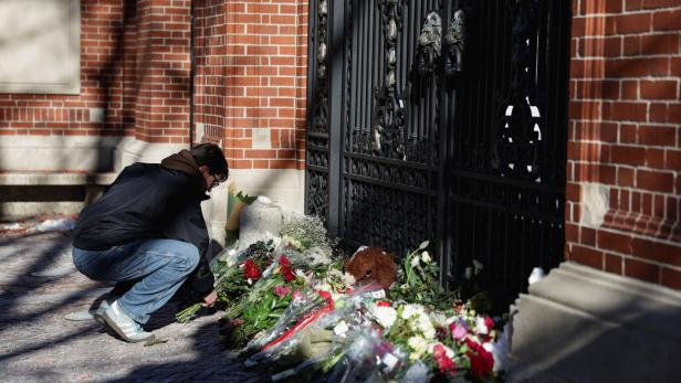 Eine Person kniet vor einem Tor und legt Blumen zu vielen bereits abgelegten Blumensträußen und einem Stofftier nieder.