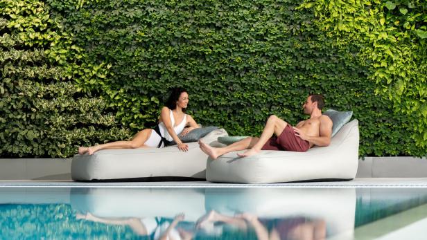 Zwei Personen entspannen auf Liegen am Pool vor einer grünen Pflanzenwand.