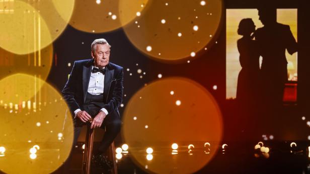 Ein älterer Mann im Smoking sitzt auf einem Hocker auf einer Bühne mit goldenen Lichtern und einer Silhouetten-Projektion im Hintergrund.