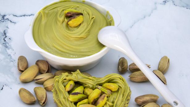 Herzförmiges Brotstück mit Pistaziencreme und Pistazien, daneben eine Schale mit Creme und ein Löffel, umgeben von Pistazien.
