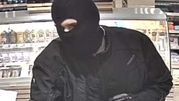 Ein maskierter Mann mit Sturmhaube, Bikerjacke hält eine Waffe in der Hand. Vor ihm steht ein Rucksack