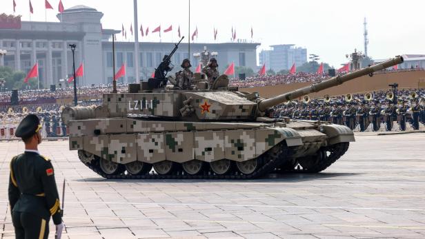 Ein Panzer mit digitaler Tarnung fährt an Soldaten und einer Menschenmenge mit roten Fahnen vorbei.