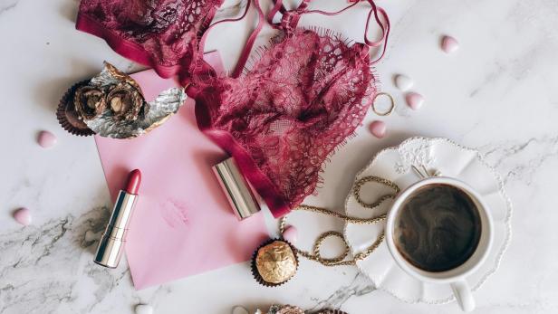 Spitzen-BH in Rot liegt neben Pralinen, Lippenstift, Schmuck und einer Tasse Kaffee auf einem Marmortisch, passend zum Silvesterbrauch für Glück im neuen Jahr.