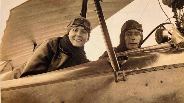 Zwei Personen mit Fliegermützen und Schutzbrillen sitzen in einem offenen Doppeldeckerflugzeug.