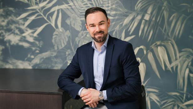 Mann im Anzug sitzt an einem Tisch vor einer Wand mit Pflanzenmuster und lächelt in die Kamera.