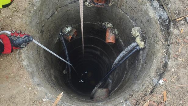 Zwei Personen in Schutzkleidung arbeiten an einem offenen, tiefen Schacht mit mehreren Rohren und Kabeln im Boden.