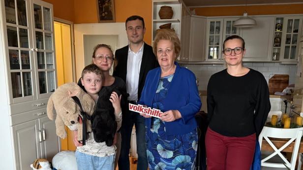 Fünf Personen stehen in einer Küche, ein Kind hält zwei Stofftiere, eine Frau hält ein Schild mit der Aufschrift „volkshilfe“.