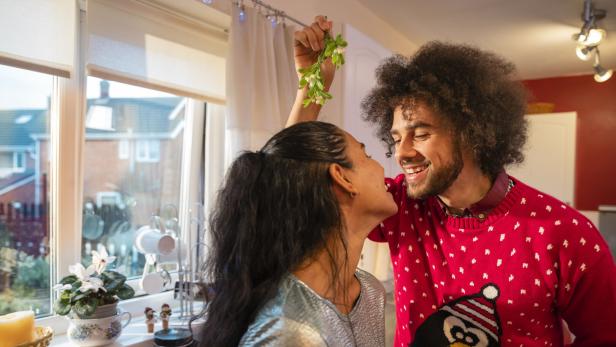 Ein Paar steht lachend in einer Küche, während eine Person Mistelzweige über die andere hält.