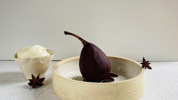 Eine pochierte Birne in einer Schale, garniert mit Sternanis, neben einem Schälchen Creme.
