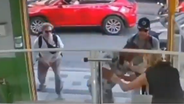 Eine Frau wird vor einem Geschäftseingang von einem Angreifer attackiert, während Passanten auf der Straße reagieren.