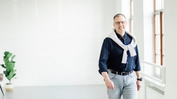 Mann mit Brille und Pullover über den Schultern steht in einem hellen, modernen Raum.