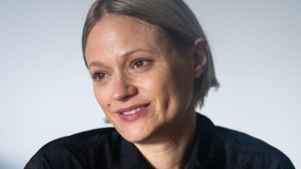 Frau mit kurzem, blondem Haar trägt eine schwarze Jacke und lächelt leicht, während sie die Arme verschränkt.