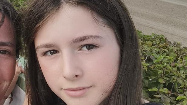 Ein Mädchen mit langen braunen Haaren steht draußen und schaut direkt in die Kamera.