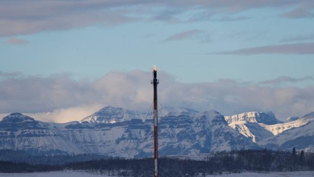 Ein hoher Schornstein mit Flamme an der Spitze steht vor verschneiten Bergen und bewölktem Himmel.
