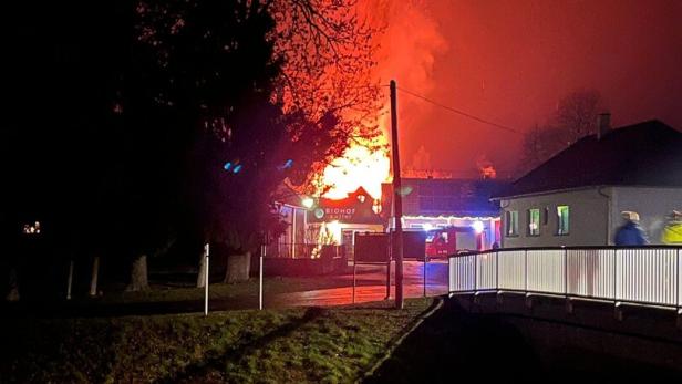 Großes Feuer mit Flammen und Funken hinter Bäumen, Einsatzfahrzeuge und Menschen in der Nähe eines Gebäudes bei Nacht.