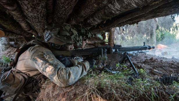 Ein Soldat in Tarnkleidung zielt mit einem Maschinengewehr aus einer getarnten Deckung im Wald.