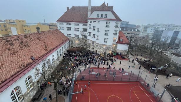 Viele Menschen versammeln sich auf einem Schulhof mit Sportplatz und historischen Gebäuden im Hintergrund.