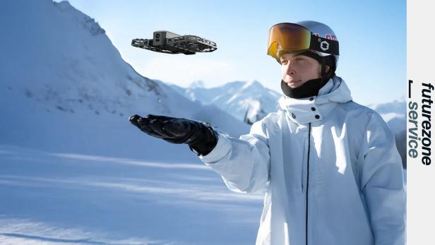 Eine Person in Winterkleidung hält eine kleine Drohne über der Hand vor verschneiten Bergen.