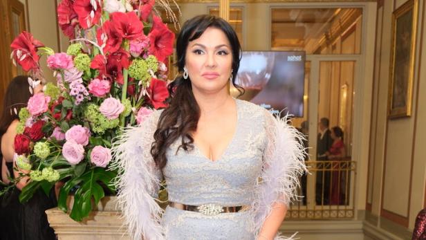 Eine Frau in einem eleganten silbernen Abendkleid mit Federn steht vor einem großen Blumenarrangement in einem prunkvollen Saal.