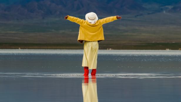 Eine Person steht mit Gummistiefeln knöcheltief in einem See, hat die Arme weit geöffnet und blickt hoffnungsvoll auf die vor ihr liegende Landschaft - und auf die Zukunft.