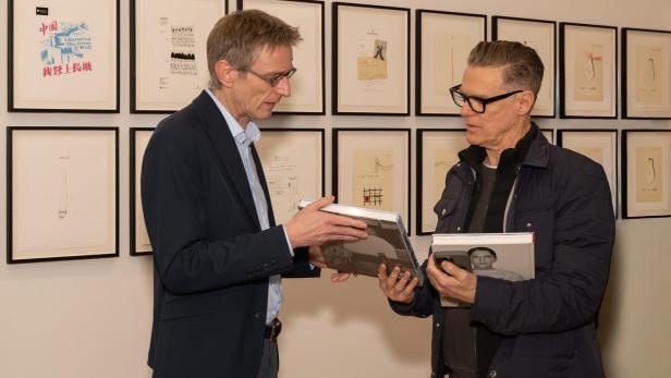 Zwei Männer unterhalten sich und halten Bücher in den Händen, im Hintergrund hängen gerahmte Zeichnungen an der Wand.