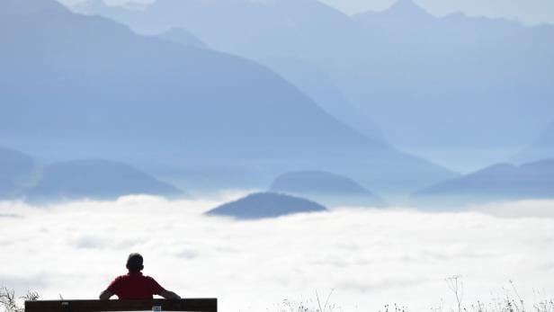 Eine Person sitzt auf einer Bank und blickt auf eine weite Berglandschaft im Nebel.