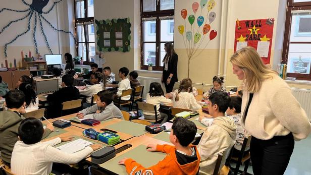 Viele Kinder sitzen an Tischen in einem Klassenraum, während zwei Erwachsene sie betreuen und unterrichten.