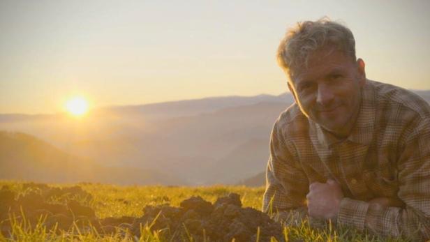 Ein Mann liegt auf einer Wiese neben einem Erdhaufen, im Hintergrund geht die Sonne über Bergen unter.
