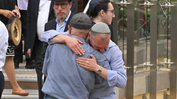 Zwei Männer umarmen sich tröstend, während andere Personen im Hintergrund stehen und ernst schauen.