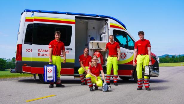 Fünf Rettungskräfte in rot-gelber Kleidung stehen und sitzen vor einem geöffneten Rettungswagen auf einer Landstraße.
