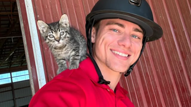 Mann in rotem T-Shirt steht mit einer Katze auf seiner Schulter und einem schwarzen Reithelm auf dem Kopf vor einer Wand