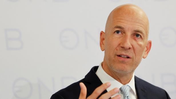 Martin Kocher spricht und gestikuliert vor einem hellen Hintergrund mit unscharfer Schrift.