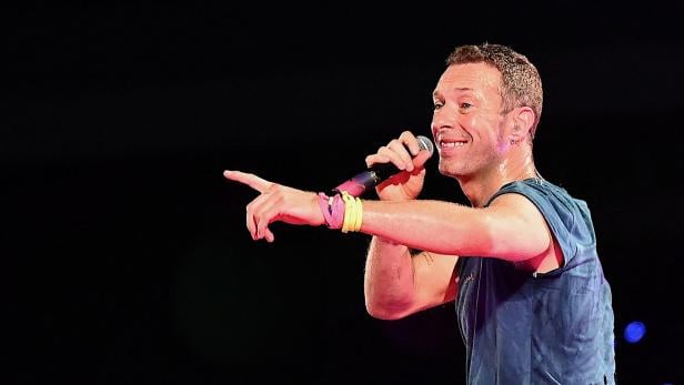 Chris Martin singt mit Mikrofon und zeigt mit dem Finger auf der Bühne.