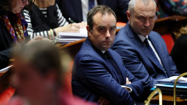 Ein Mann im blauen Anzug sitzt mit verschränkten Armen neben anderen Personen.